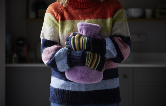 Eine Person mit gestreiftem Pullover und Armstulpen umklammert auf Bauchhöhe eine Wärmflasche.