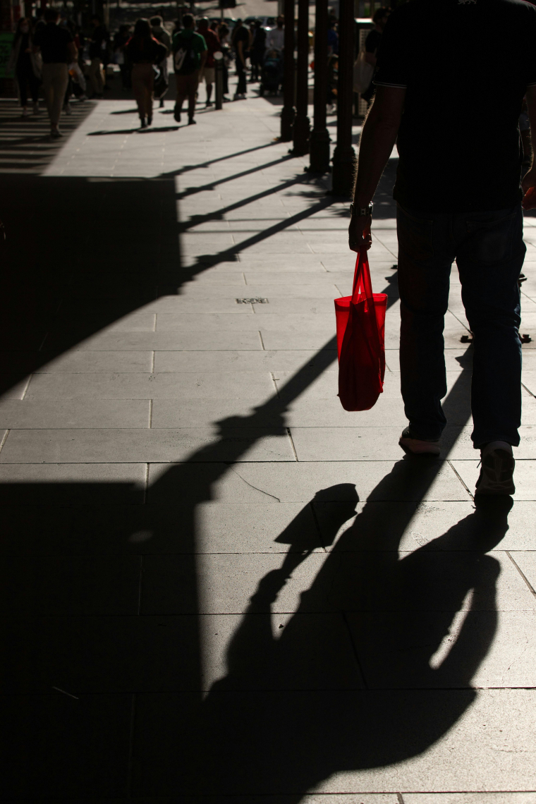 Person mit roter Einkaufstasche, von hinten fotografiert, Schatten von Laternen und der Person auf Betonboden sichtbar