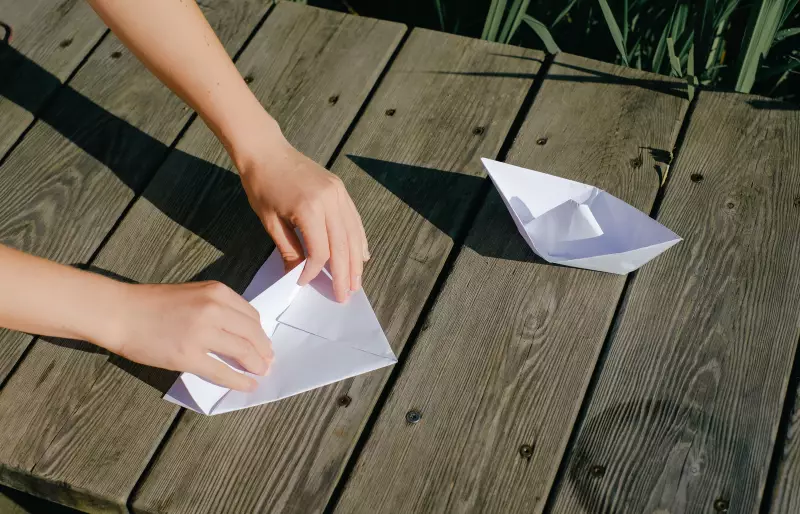 Zwei Hände falten ein Papierboot. Ein zweites Papierboot liegt fertig daneben.