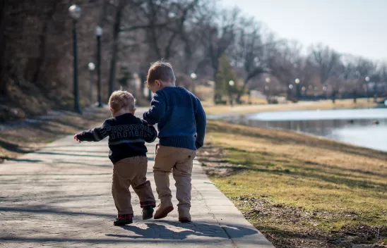 Zwei Kinder stehen auf einer Promenade, man sieht sie von hinten und sie halten sich an den Händen. Rechts davon ist eine Wiese und ein See zu sehen.
