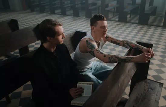 Ein Priester mit einer Bibel im Schoß sitzt neben einem Mann mit Tanktop und Tattoos auf einer Kirchenbank in einer leeren Kirche. 