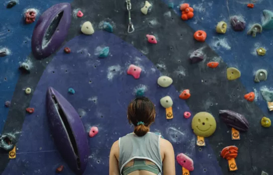 Eine Frau steht vor einer blauen Kletterwand mit bunten Klettergriffen und schaut nach oben. Eine Frau steht vor einer blauen Kletterwand mit bunten Klettergriffen und schaut nach oben.