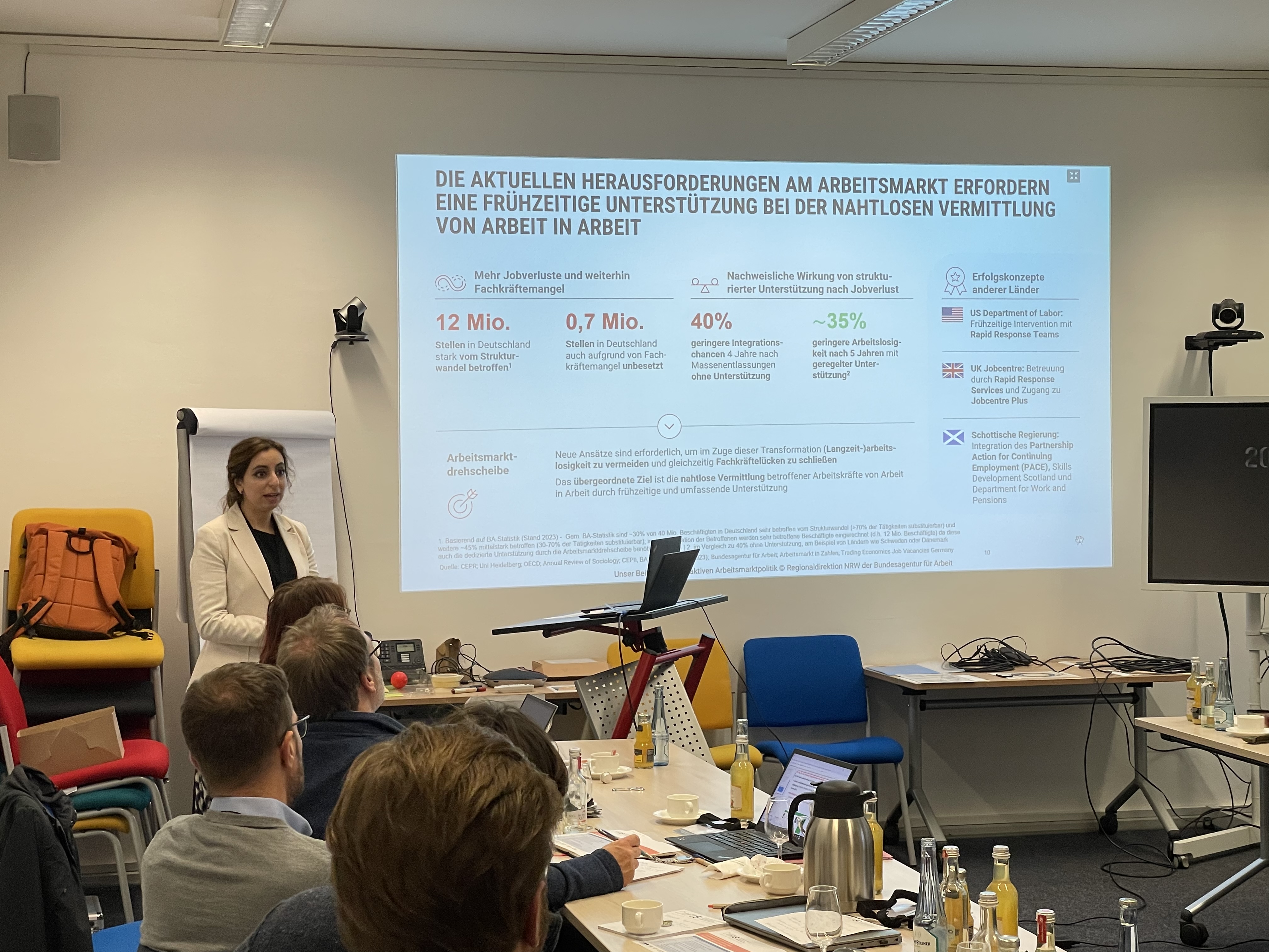 Eine Person steht vor einer Leinwand, auf der eine Präsentation läuft und hält einen Vortrag vor Publikum