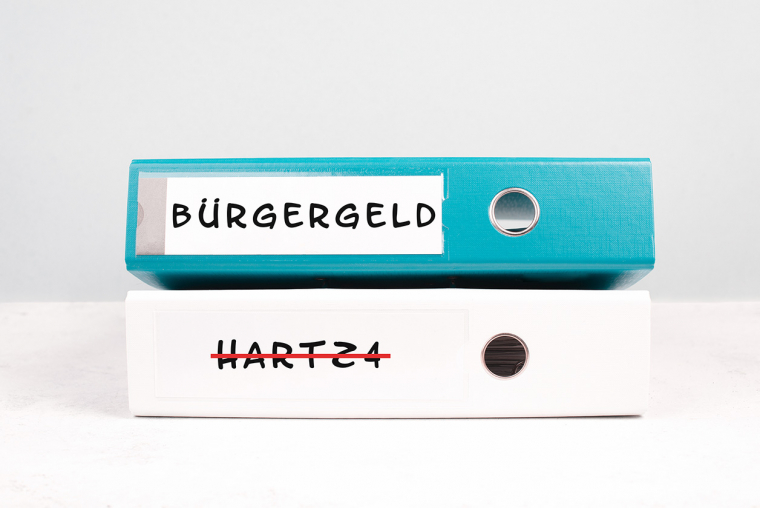 Ein blauer Ringordner mit der seitlichen Aufschrift "Bürgergeld" liegt auf einem weißen Ringordner mit der in rot durchgestrichenen Aufschrift "Hartz 4".