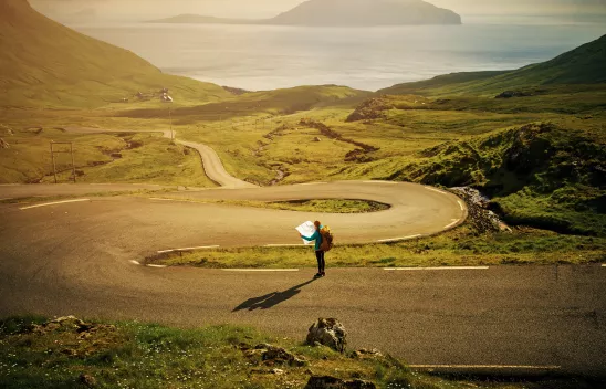 Eine Person mit einer Landkarte in den Händen steht auf einer Serpentinenstraße zwischen grünen Wiesen, die am Meer mündet. Eine Person mit einer Landkarte in den Händen steht auf einer Serpentinenstraße zwischen grünen Wiesen, die am Meer mündet.