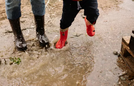 Beine zweier Personen, eine erwachsen, eine im Kindesalter, mit Gummistiefeln bekleidet in einer Pfütze springend. 