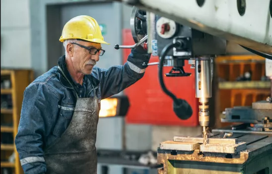 Ein älterer Arbeiter mit Schutzkleidung und gelbem Schutzhelm arbeitet an einer Metallschneidemaschine in einer Halle. Ein älterer Arbeiter mit Schutzkleidung und gelbem Schutzhelm arbeitet an einer Metallschneidemaschine in einer Halle.