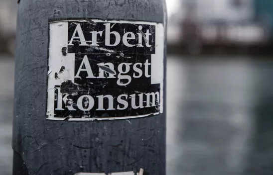 Ein zerkratzter Sticker mit der Aufschrift "Arbeit, Angst, Konsum" klebt an einer Laterne. Das Foto ist Schwarzweiß. 