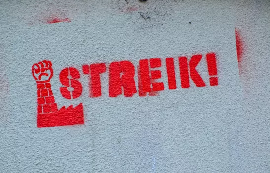 Ein rotes Graffiti an einer hellen Wand zeigt den Schriftzug "Streik!" und eine erhobene Faust. Ein rotes Graffiti an einer hellen Wand zeigt den Schriftzug "Streik!" und eine erhobene Faust.